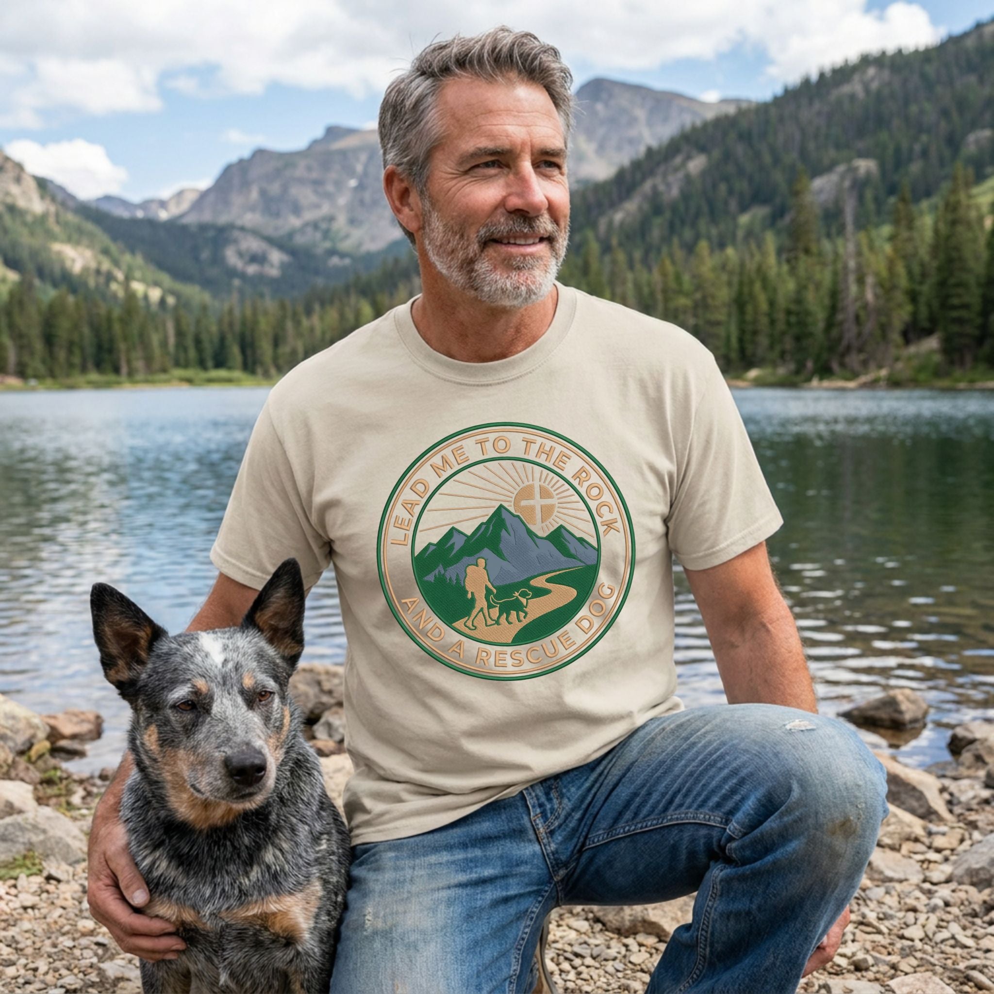 Man in a beige "Lead Me To The Rock" graphic tee with a rescue dog by a mountain lake.