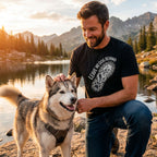 Man in black "Leave No Dog Behind" tee petting a husky by a lake at sunset.