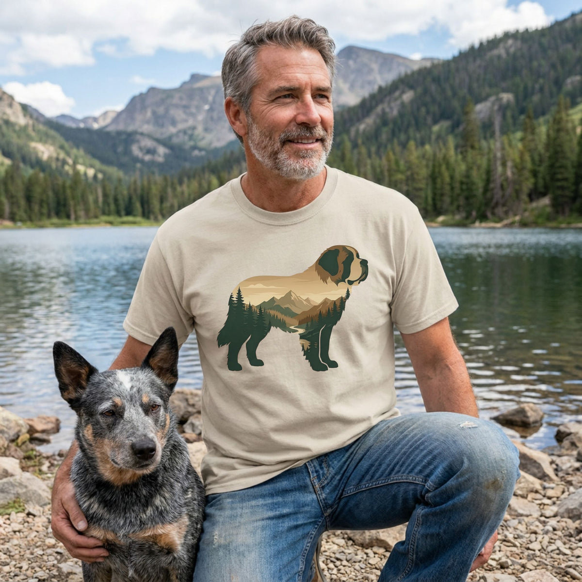 Man in a tan Mountain Dog T-Shirt with a scenic dog graphic, kneeling beside a blue heeler by a lake.