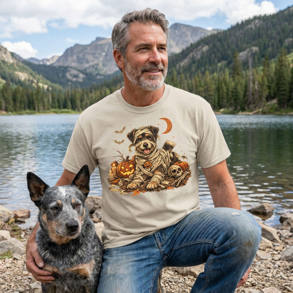 Man wearing a beige t-shirt with a mummy dog, jack-o'-lanterns, and skull graphic, posing with a dog by a mountain lake.
