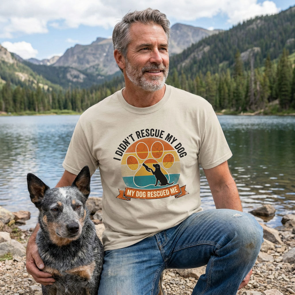 Man in a beige "I Didn't Rescue My Dog, My Dog Rescued Me" tee with a blue heeler by a lake.