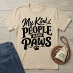 Beige crewneck t-shirt with My Kind of People Have Paws text, displayed with blue jeans and a dog treat bag on wood.