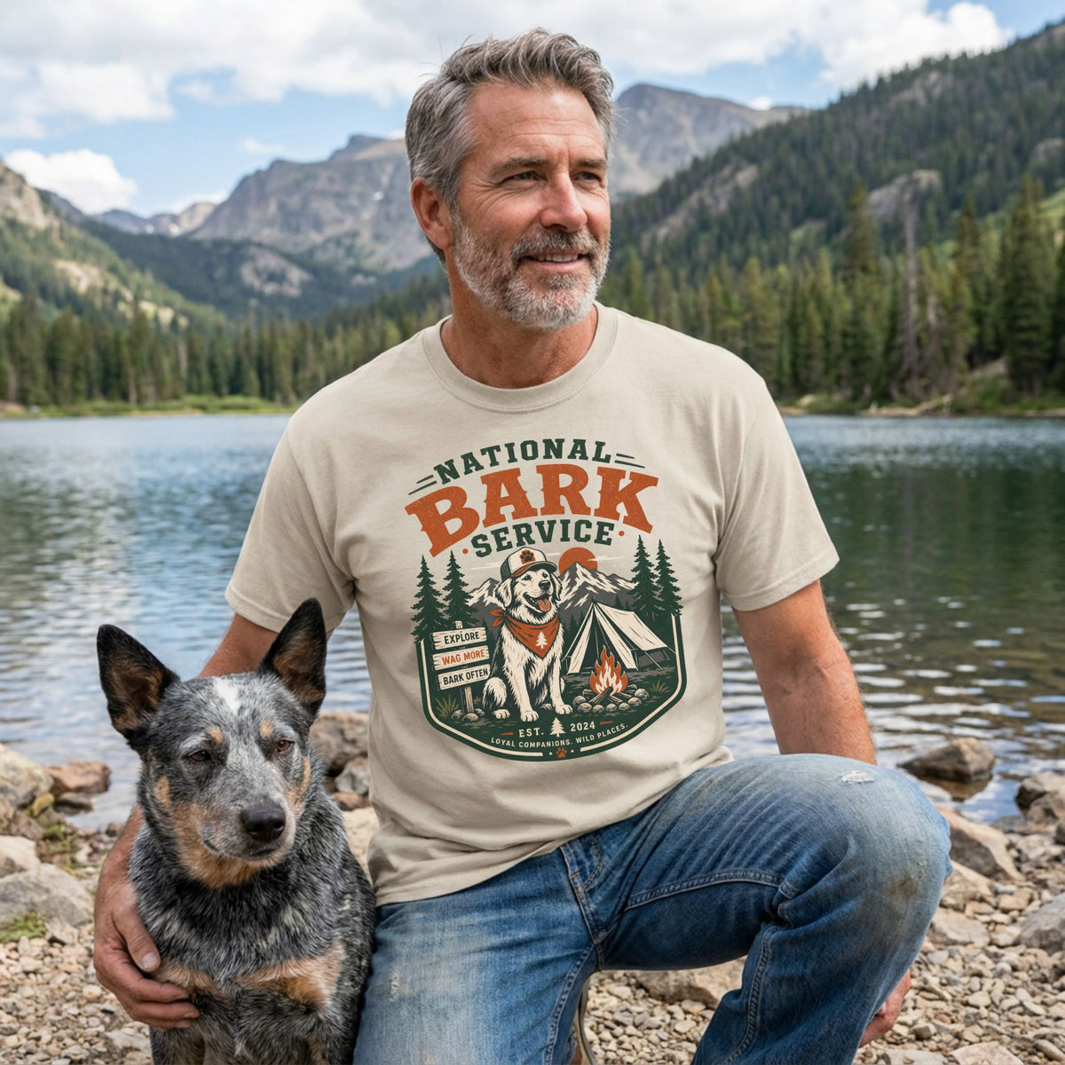 Man wearing a cream-colored National Bark Service graphic t-shirt featuring a dog, tent, and mountain scene by a lake.