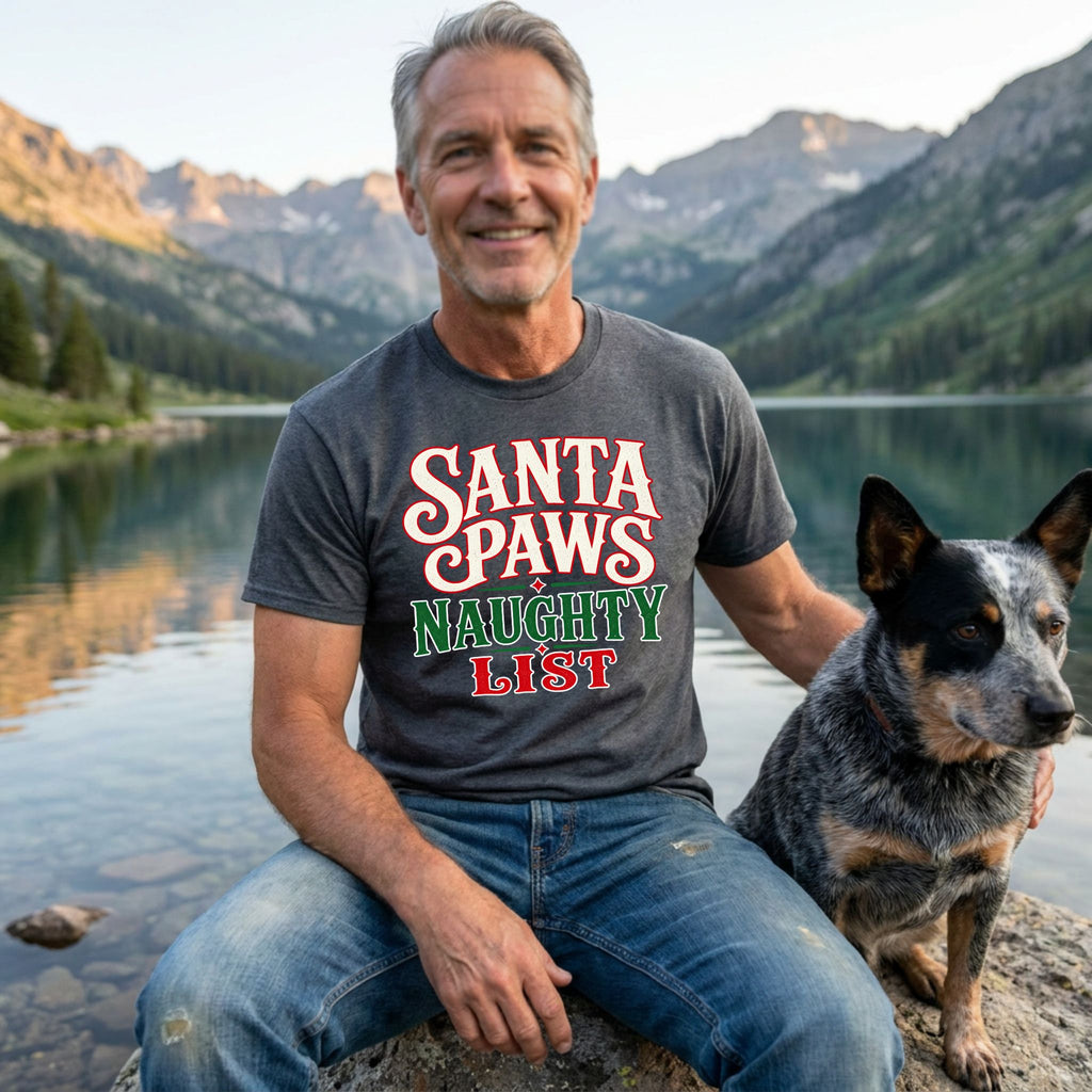 Man wearing a charcoal grey T-shirt with Santa Paws Naughty List text in red, green, and white, sitting by a mountain lake.
