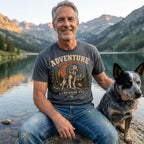 Man wearing a charcoal grey graphic t-shirt featuring a dog hiking illustration, sitting by a mountain lake with his dog.