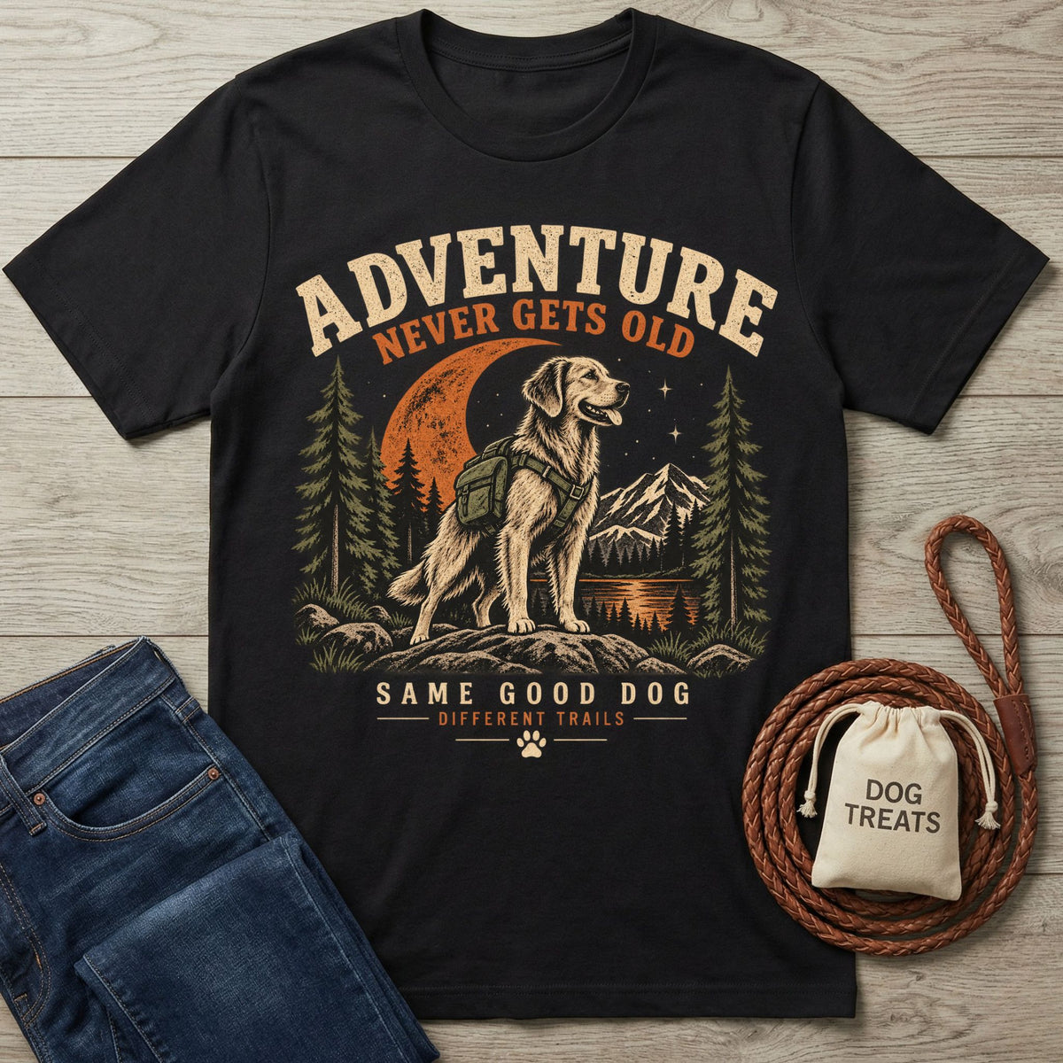 Black crewneck t-shirt featuring a golden retriever hiking graphic with mountains, trees, and the text Adventure Never Gets Old.