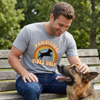 Man in a grey "Pawsitive Vibes Only" t-shirt petting a German Shepherd on a park bench.