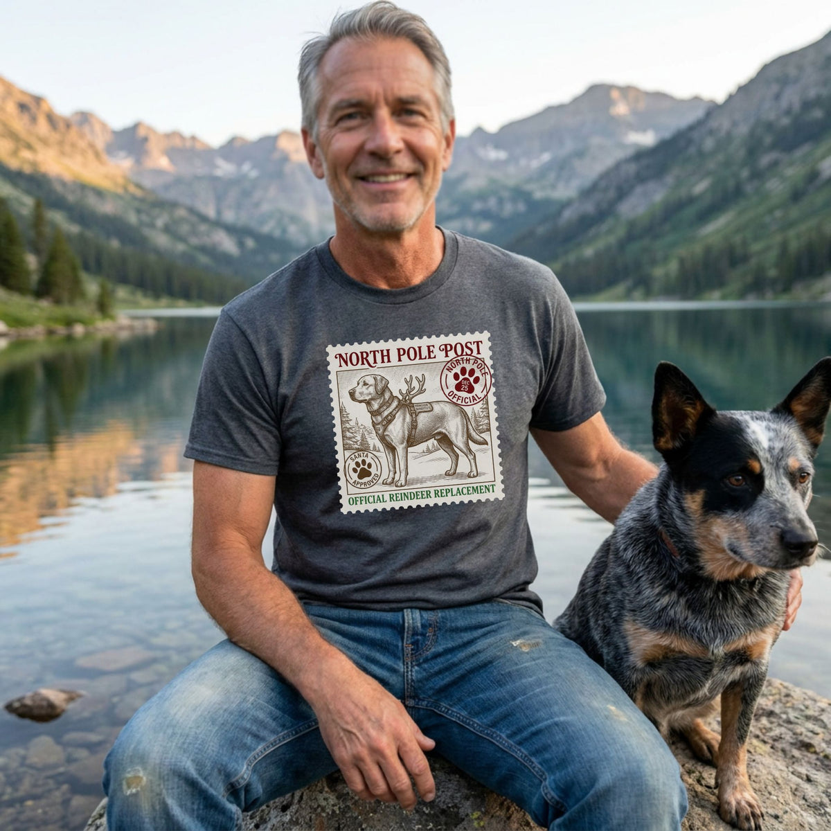 Man in a charcoal grey "Official Reindeer Replacement" dog graphic tee, blue jeans, with a blue heeler dog by a lake.