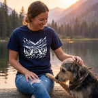 Woman in navy Rescue Ridge T-shirt with mountain and dog graphic, petting a dog by a lake at sunset.