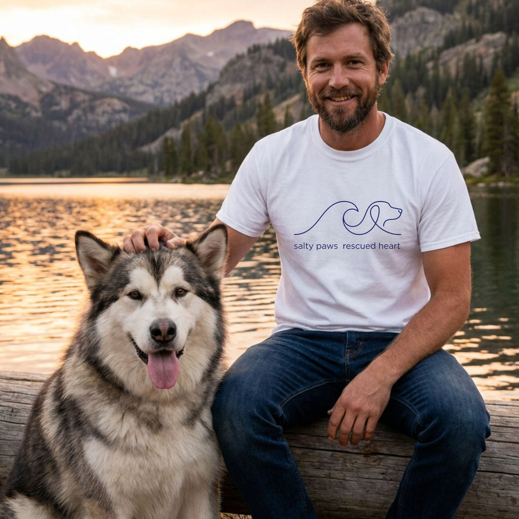 Man in white "Salty Paws Rescued Heart" tee with a Malamute by a lake at sunset.