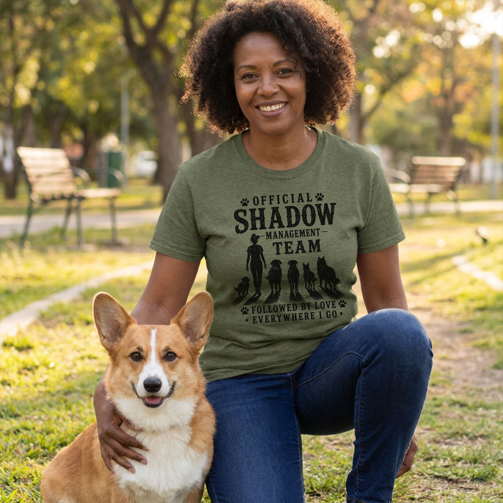 Woman in an olive green "Shadow Management Team" t-shirt, kneeling with a Corgi in a park.