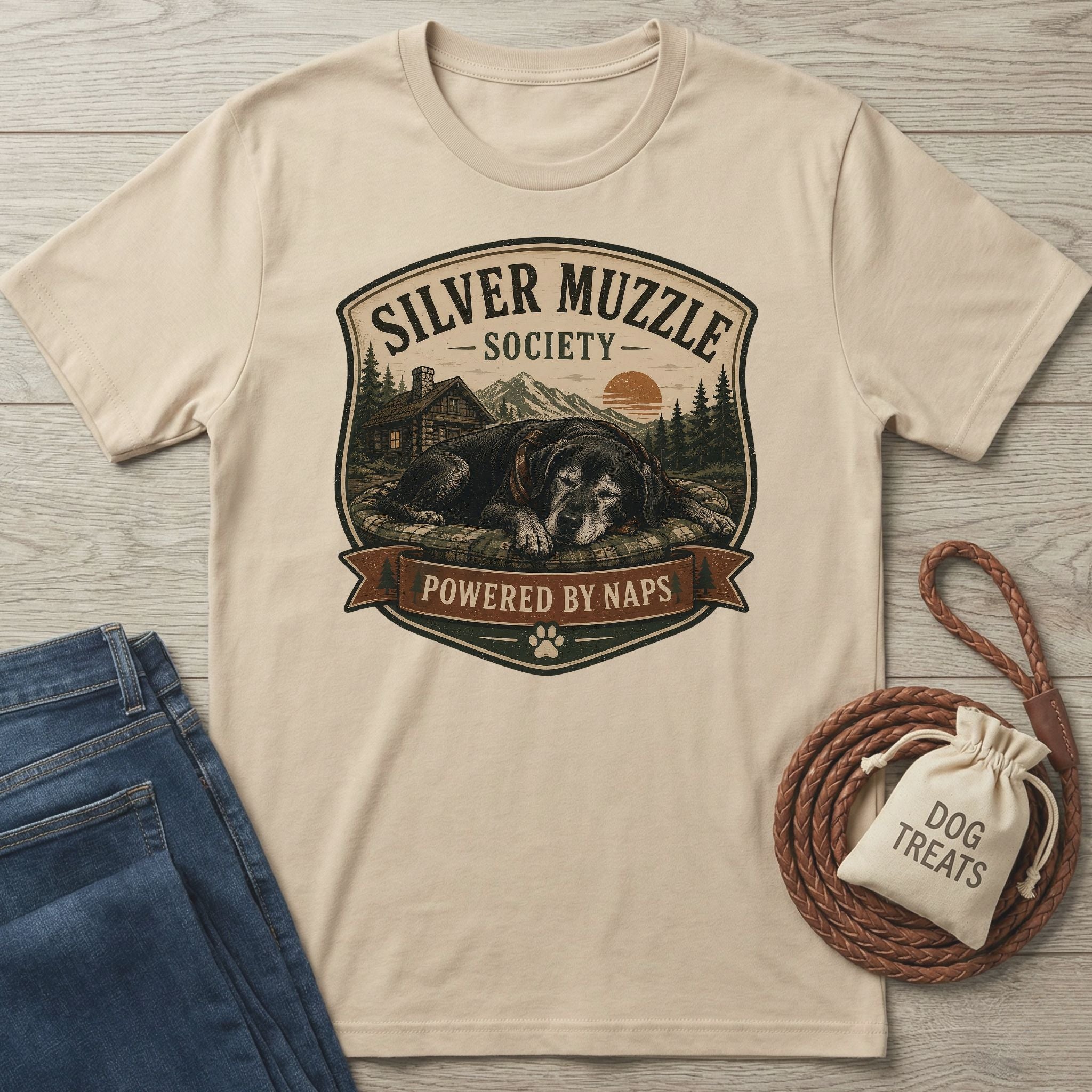 Beige "Silver Muzzle Society" t-shirt with a sleeping senior dog graphic, next to jeans and dog treats.