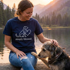 Woman in a navy "simply blessed" dog and heart graphic tee petting a scruffy dog by a lake at sunset.