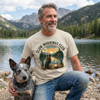 Man wearing a beige Slow Mornings Club graphic t-shirt featuring a dog and lakeside sunrise, kneeling beside his dog.