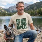 Man in a light tan "Stay Home. Pet Dogs. Repeat." t-shirt with a blue heeler dog by a lake.