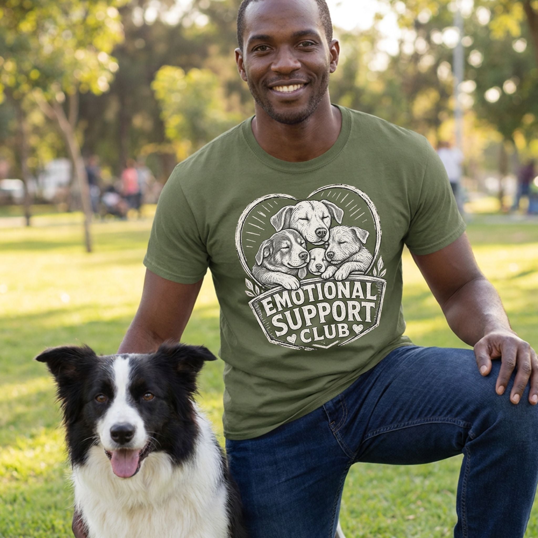 Man wearing an olive green t-shirt with a white graphic of three puppies and the text Emotional Support Club in a park.