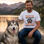 Man in a white "Saving The Old Guard" senior dog rescue t-shirt petting a large fluffy dog by a lake at sunset.