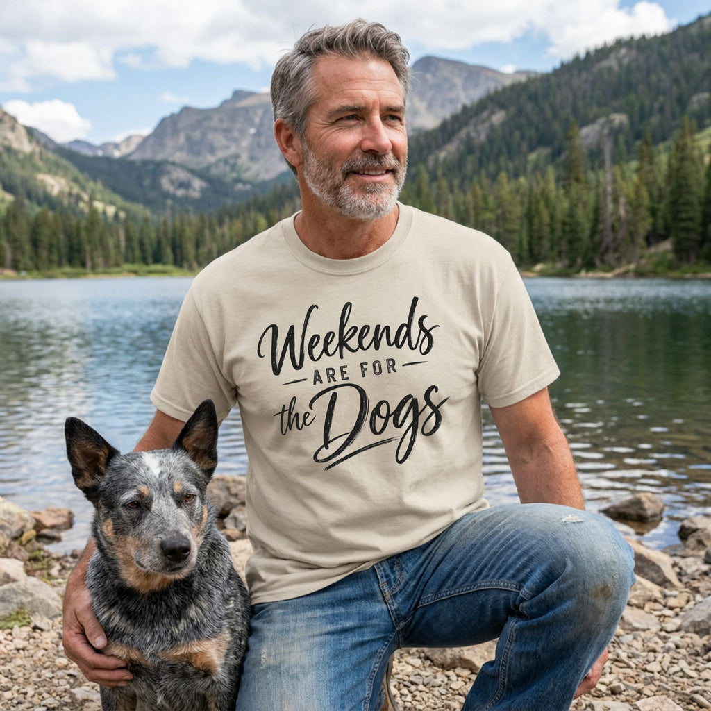 Man wearing a beige crewneck T-shirt with Weekends Are For The Dogs text, kneeling by a lake with his dog.