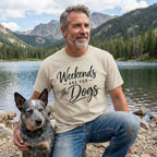 Man wearing a beige crewneck T-shirt with Weekends Are For The Dogs text, kneeling by a lake with his dog.