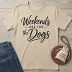 Beige crewneck t-shirt with black script text reading Weekends Are For The Dogs, laid flat with jeans and a treat bag.