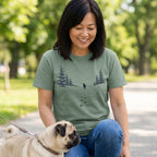 Woman wearing a sage green cotton t-shirt with a mountain, pine forest, and dog paw print graphic while walking a pug.