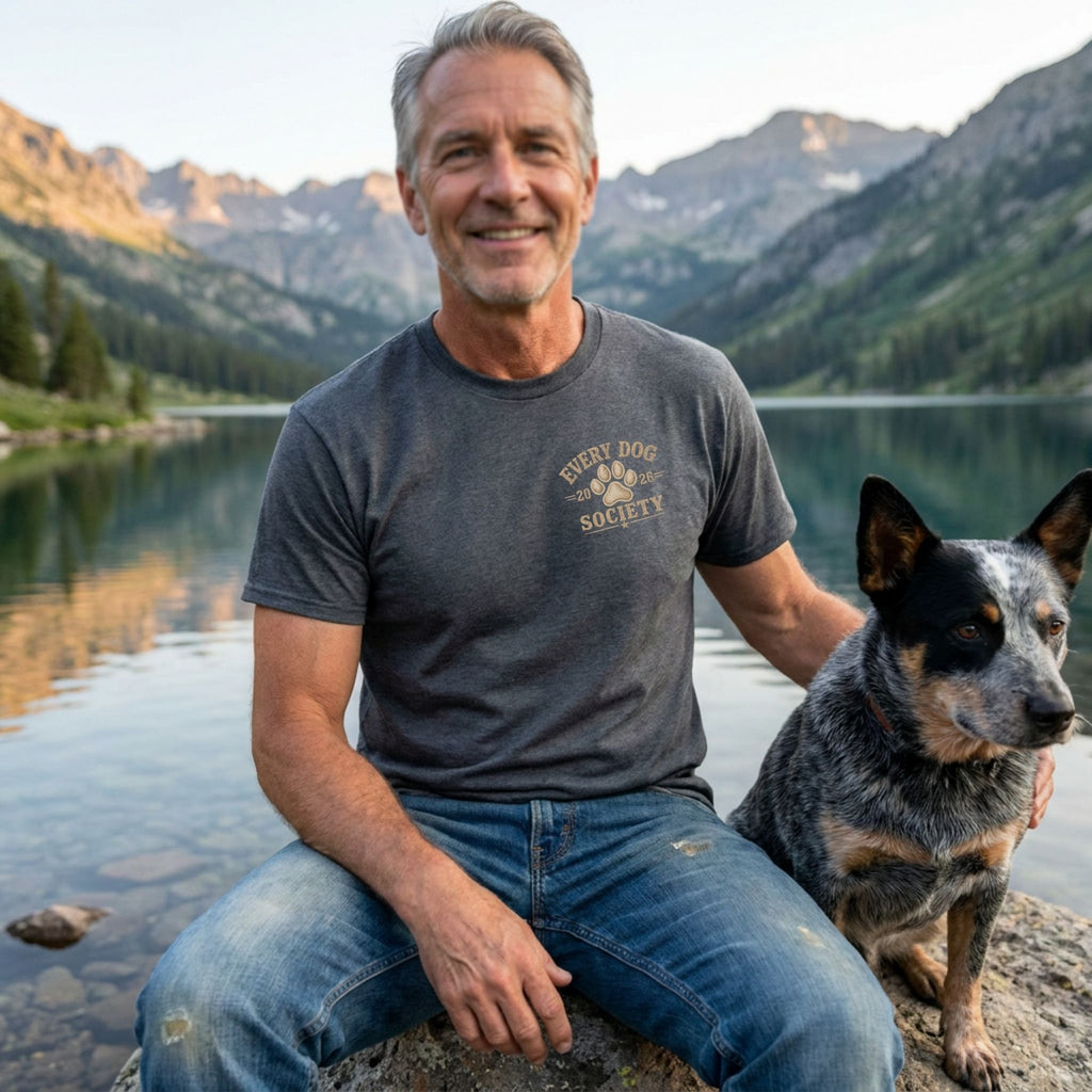 Man wearing a charcoal grey vintage-style World Tour t-shirt with a paw print logo, sitting by a lake with his dog.