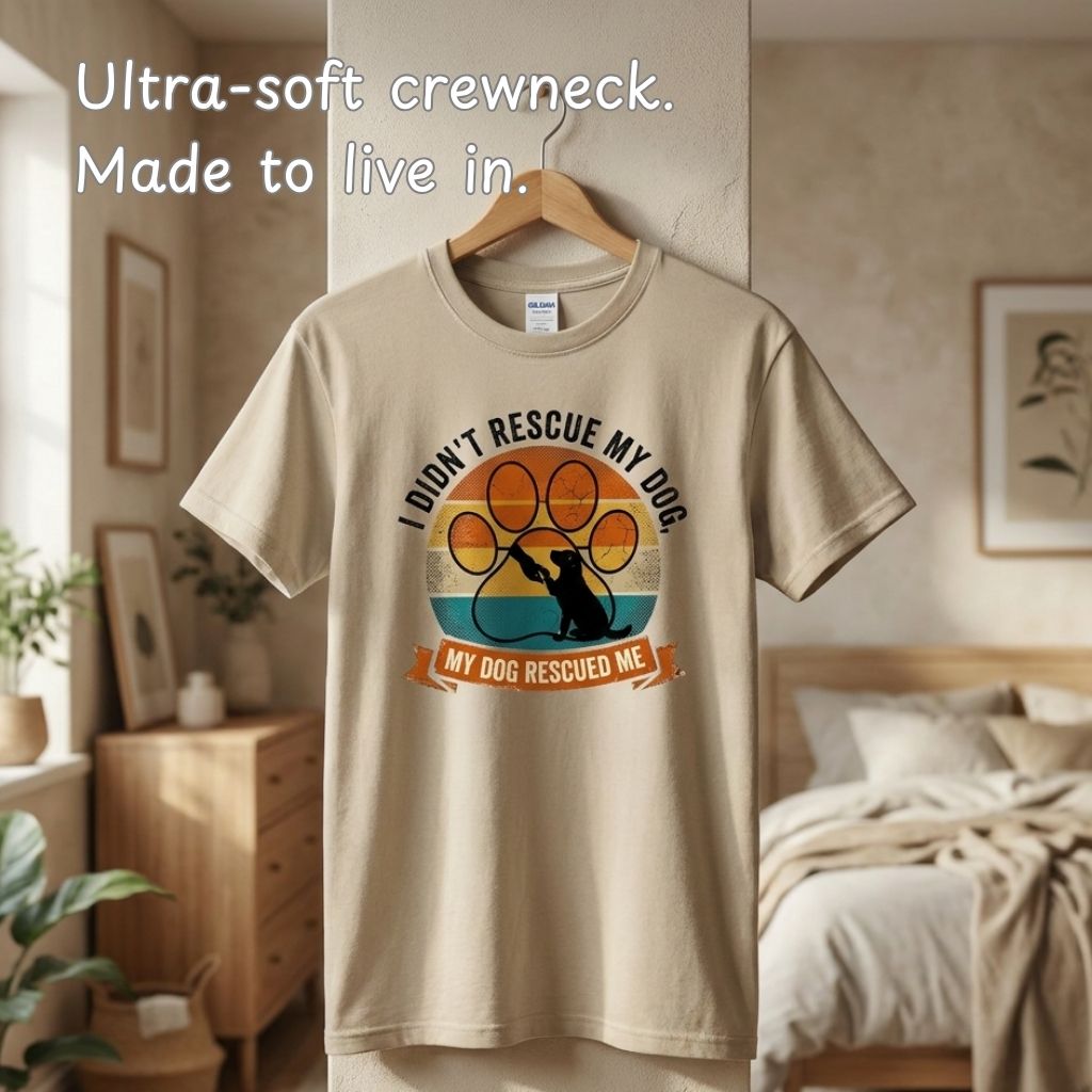 A beige crewneck T-shirt hanging on a wooden hanger in a softly lit bedroom. The shirt has a colorful graphic with a dog silhouette over a paw print and the text 'I DIDN'T RESCUE MY DOG.